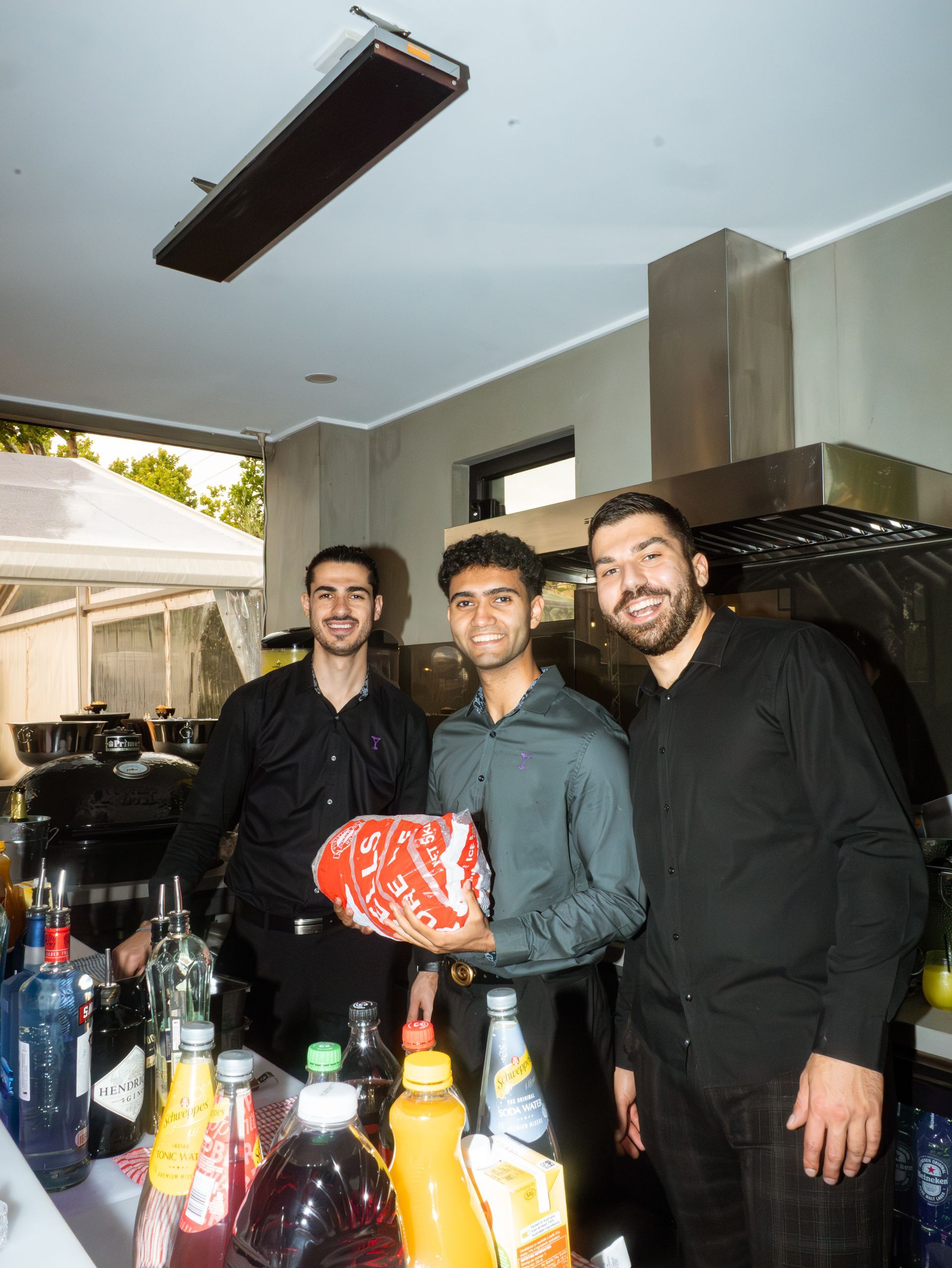 Three men standing behind a table of drinks at an outdoor gathering, smiling at the camera.