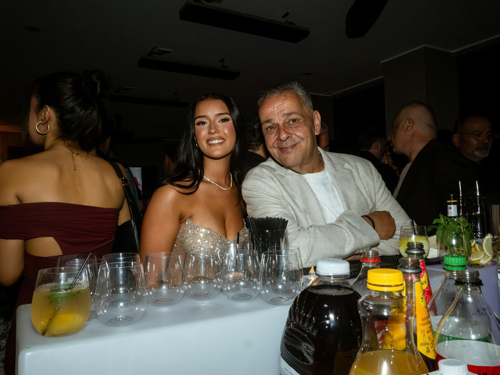 Two people smiling at a dimly lit bar table with drinks and bottles in front of them