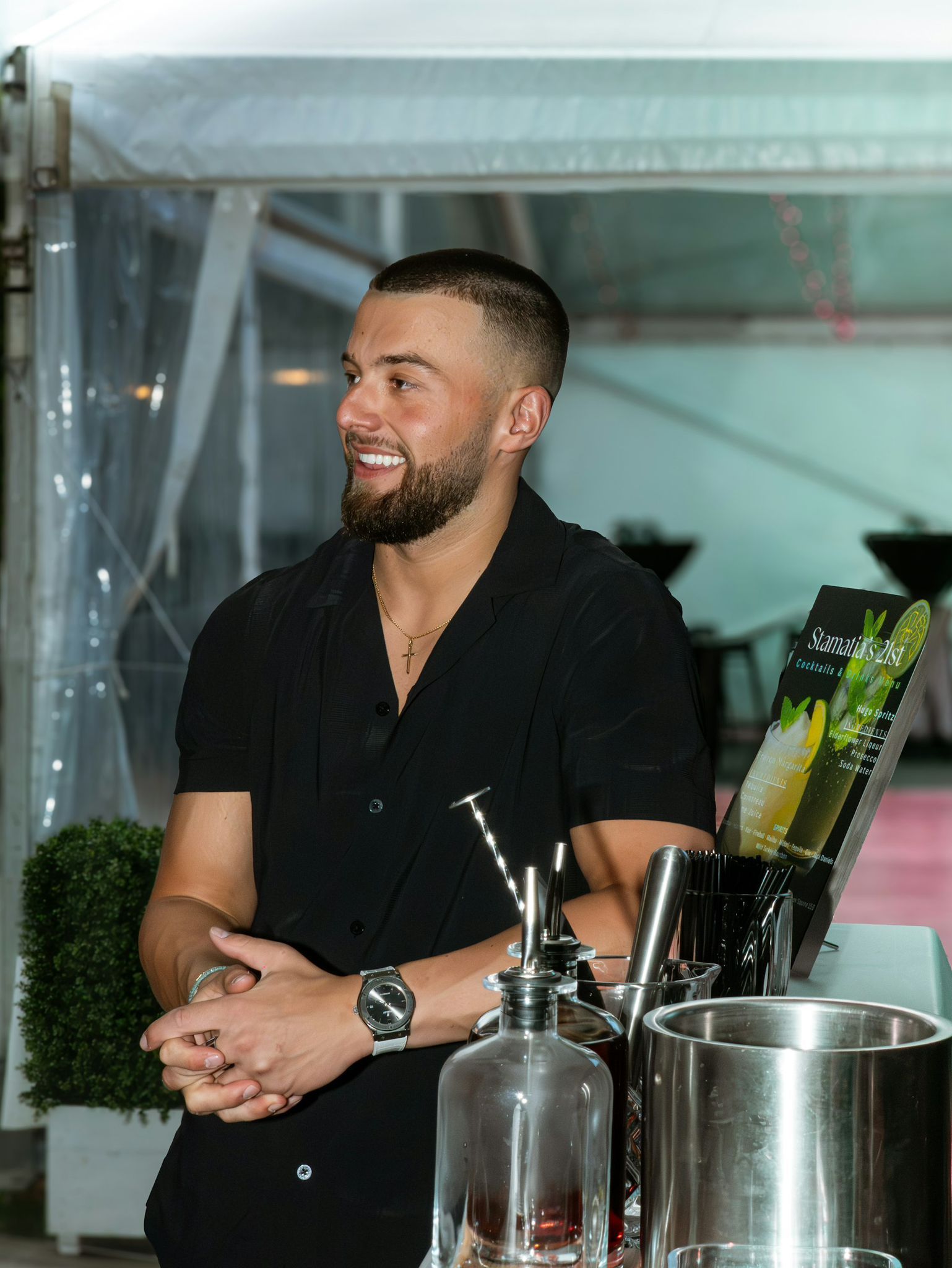 Man smiling behind a bar, wearing a black shirt, with bottles and glassware in the foreground
