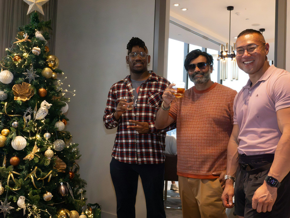 Three people stand beside a decorated Christmas tree, smiling and holding drinks in a hallway.