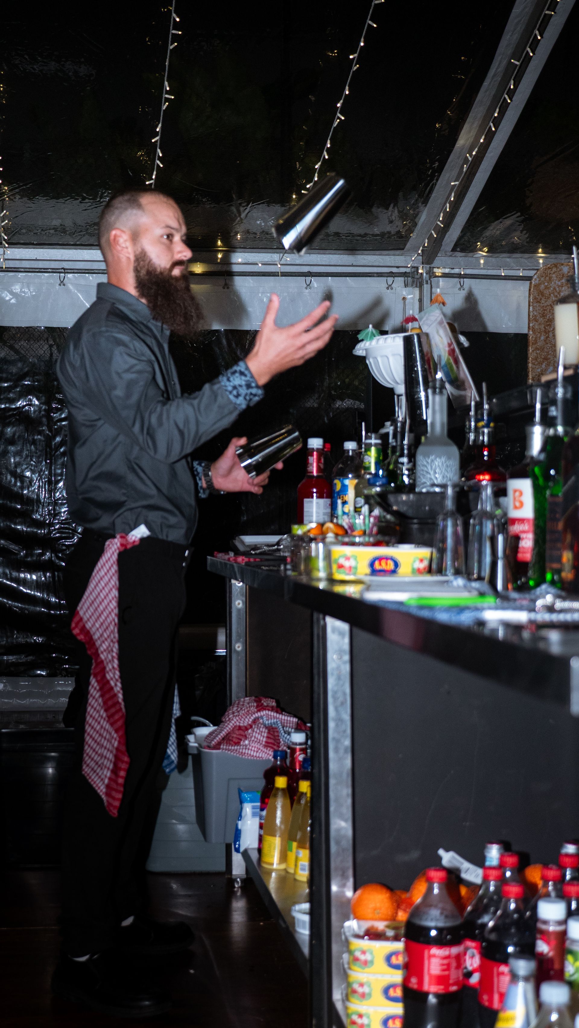 Bartender in a dark bar, gesturing near a crowded counter with bottles and drink mixers.