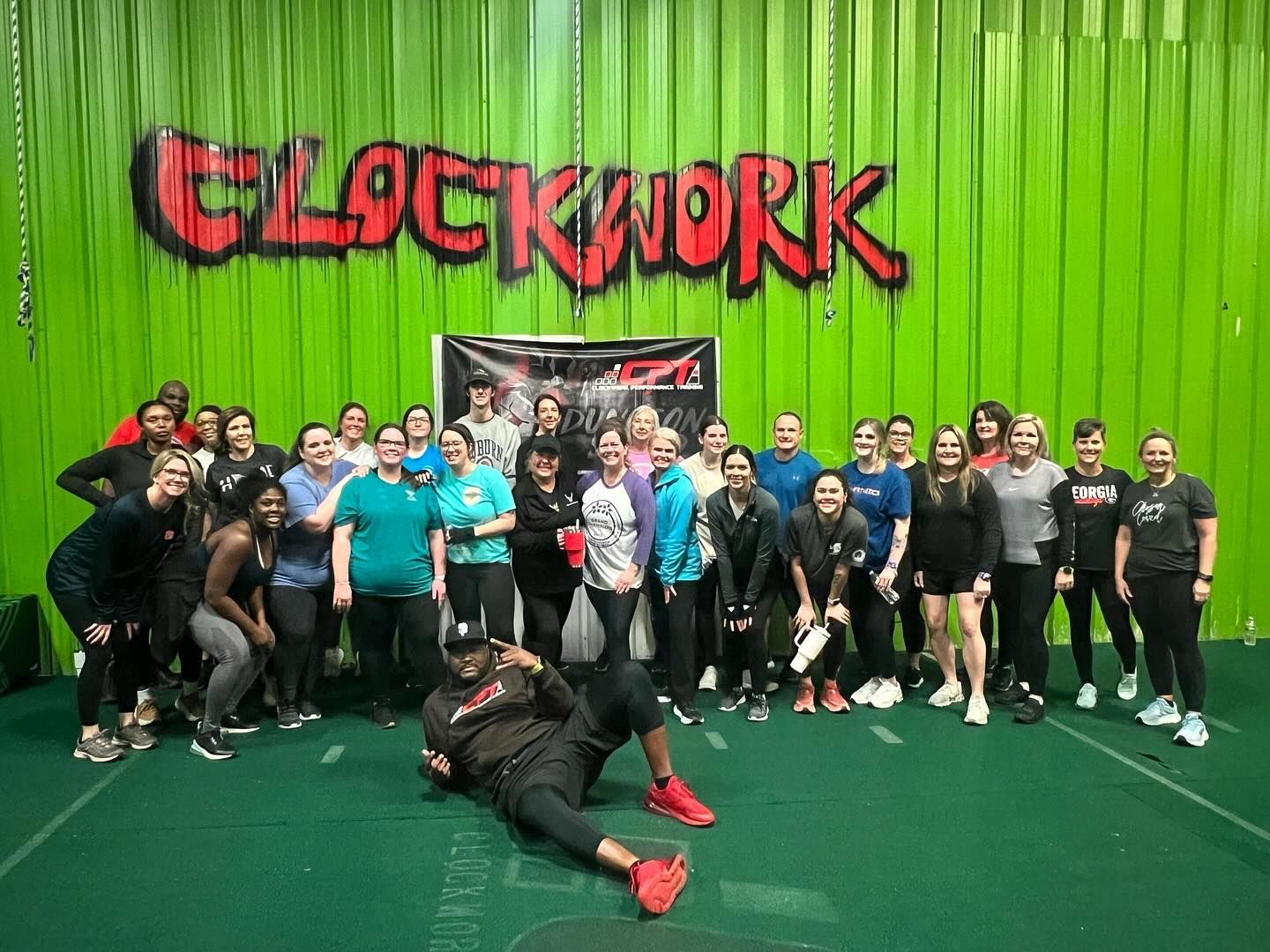 Group of people posing in a gym under a red-and-black “Clockwork” banner on a green wall
