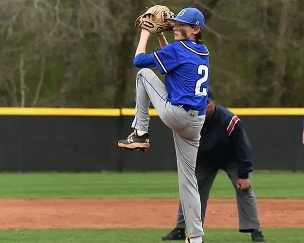 Baseball pitcher in blue jersey and gray pants winds up on the mound, mid-throw with glove raised.