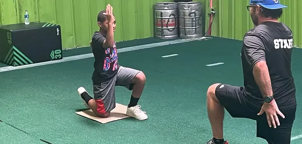 Two athletes kneel on turf during a training drill near a coach in a blue cap.