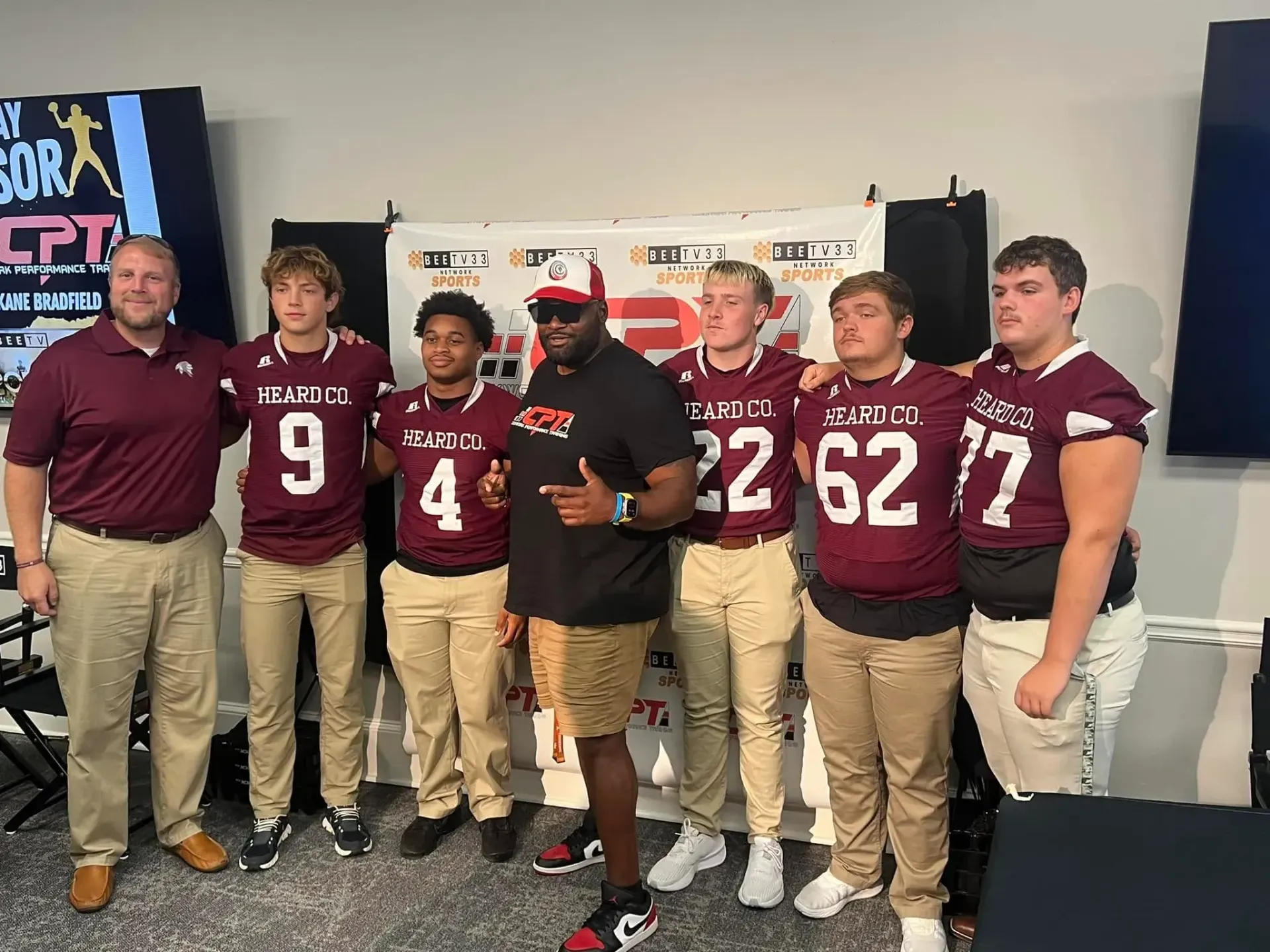 Six football players and a coach pose indoors in maroon and tan uniforms at a team event