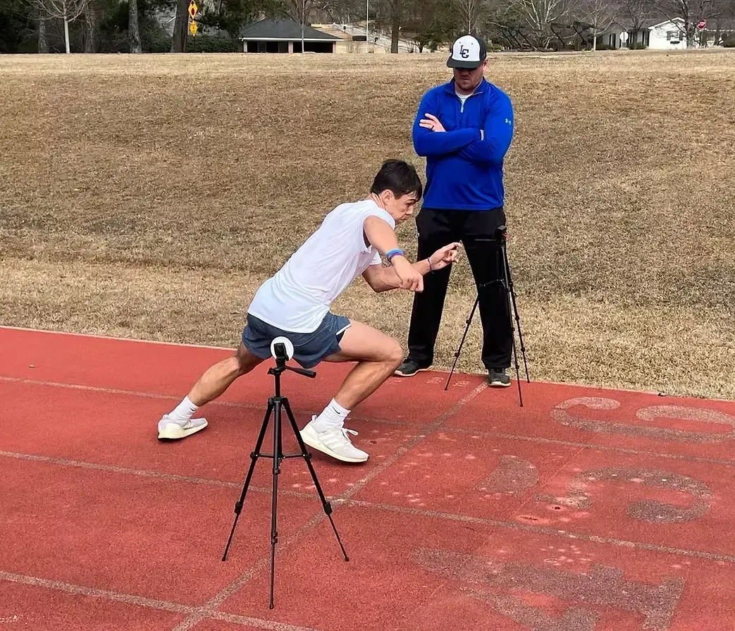 Athlete lunging on a track while coach observes beside a tripod.