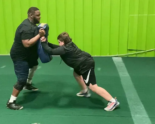 Two people wrestling on green mats in a gym, one holding focus pads for training.