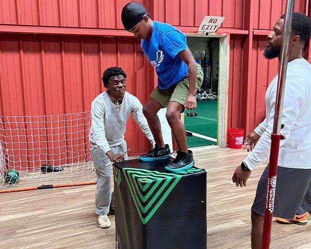Man balancing on a padded box while two people spot him in an indoor gym.