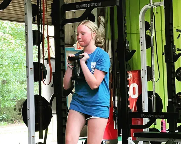 Person exercising with a kettlebell in a gym, wearing a blue shirt and shorts.