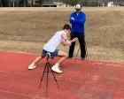 Athlete in white shirt lunging on a track while coach watches beside him, with timing tripod in front.