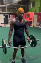 Man lifting a hex bar in a gym, wearing a yellow head covering and black workout clothes.
