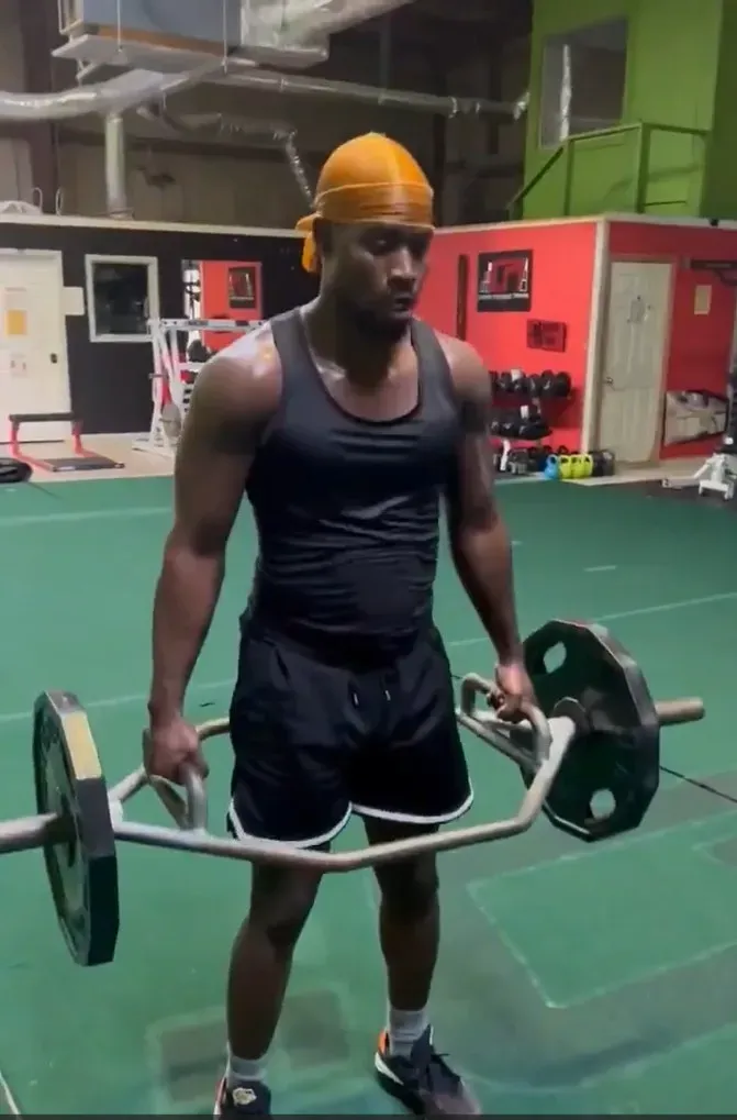 Man lifting a hex bar in a gym, wearing a yellow head covering and black workout clothes.
