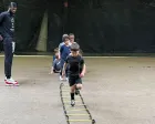 Children doing ladder drills on an outdoor field while a coach watches nearby
