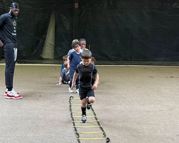 Children doing ladder drills on an outdoor field while a coach watches nearby