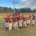 Youth baseball team posing on a field at sunset, wearing orange and white uniforms, with trees in the background