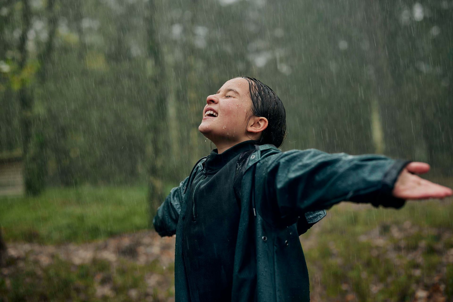 Een persoon met uitgestrekte armen geniet van de regen in een bosrijke omgeving.