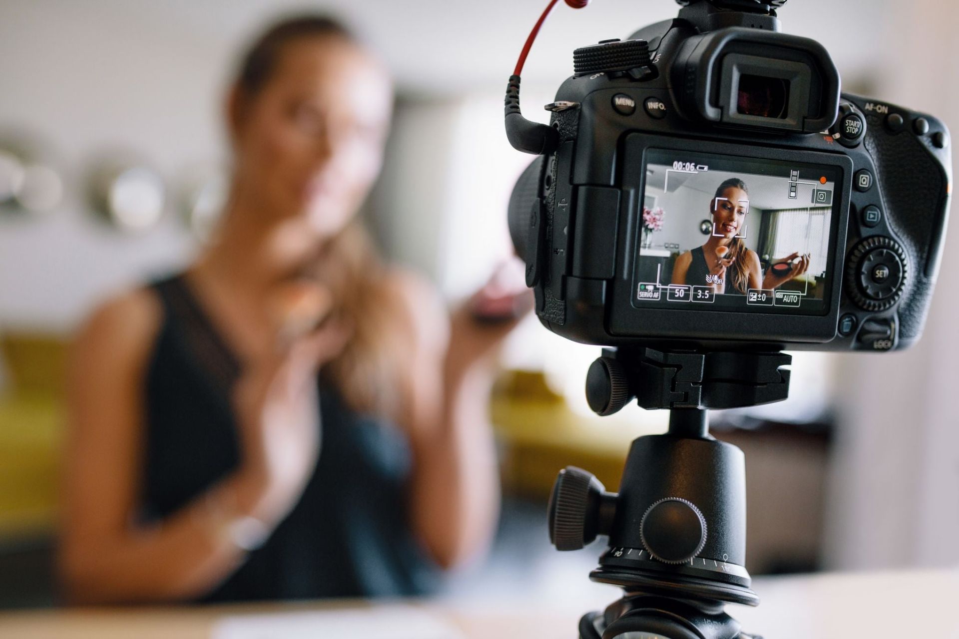 Een camera op statief filmt een vrouw binnenshuis; op het camerascherm is te zien hoe de vrouw gebaren maakt, tegen een wazige achtergrond.