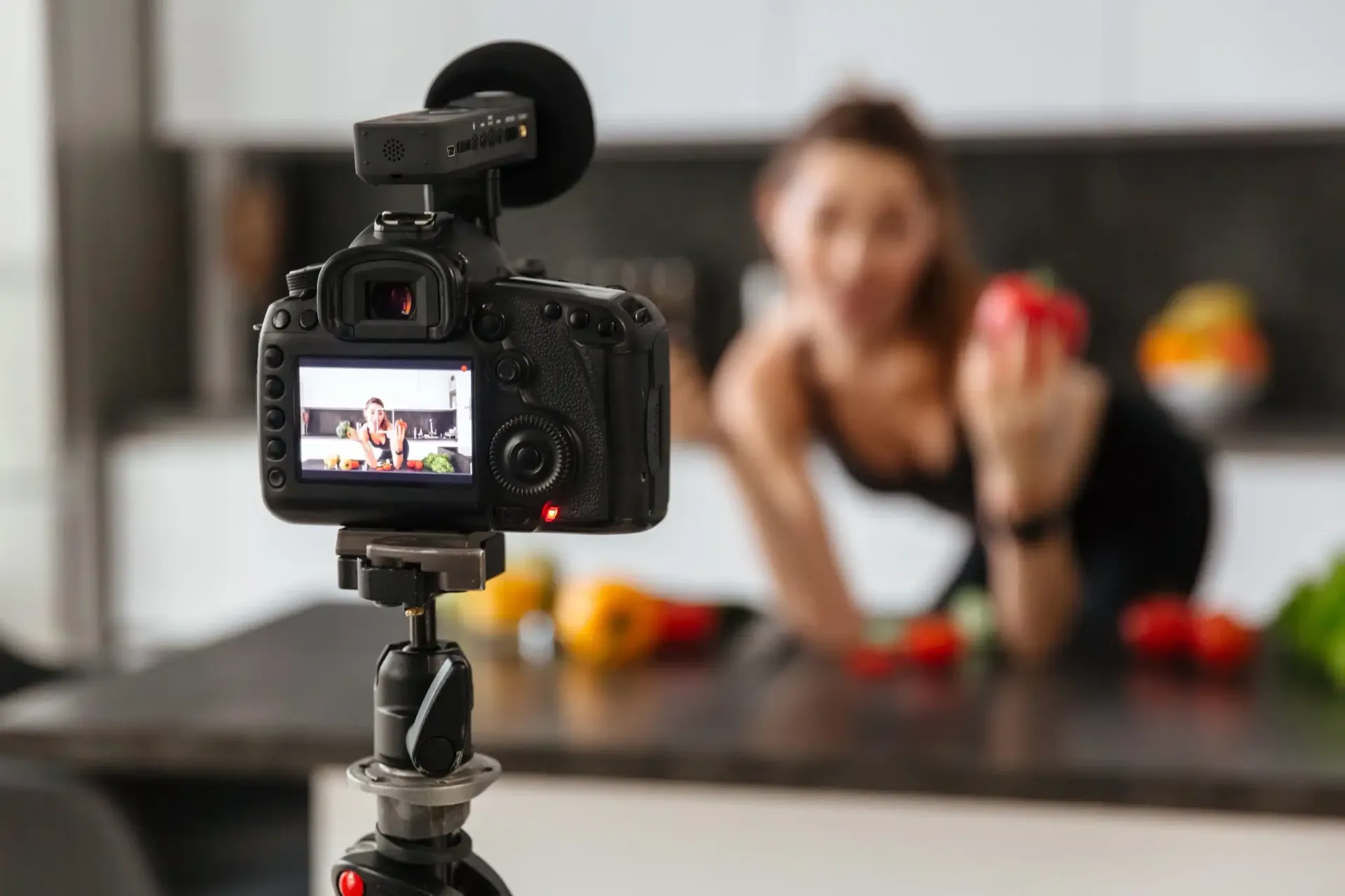 Een camera filmt een vrouw in de keuken die een rode paprika vasthoudt. Groenten liggen op het aanrecht.