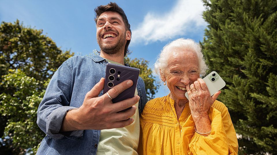 Een man en een oudere vrouw glimlachen en houden smartphones vast, buiten bij bomen onder een blauwe hemel.