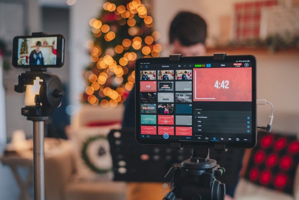 Tablet en telefoon op statieven tonen live video; persoon op de achtergrond met kerstboom.