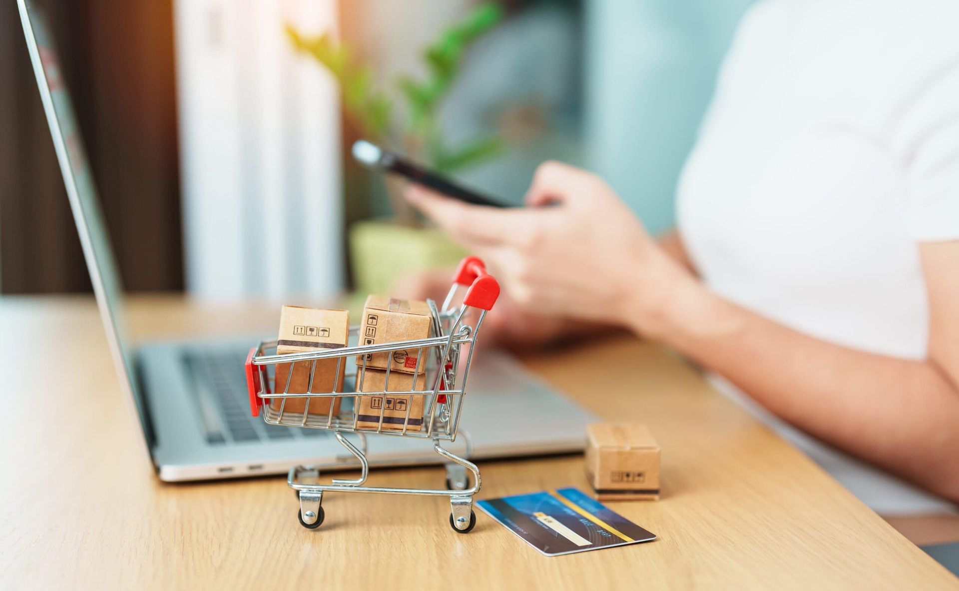 Een persoon die online winkelt met een laptop en telefoon, een klein winkelwagentje met dozen en een creditcard op een houten bureau.