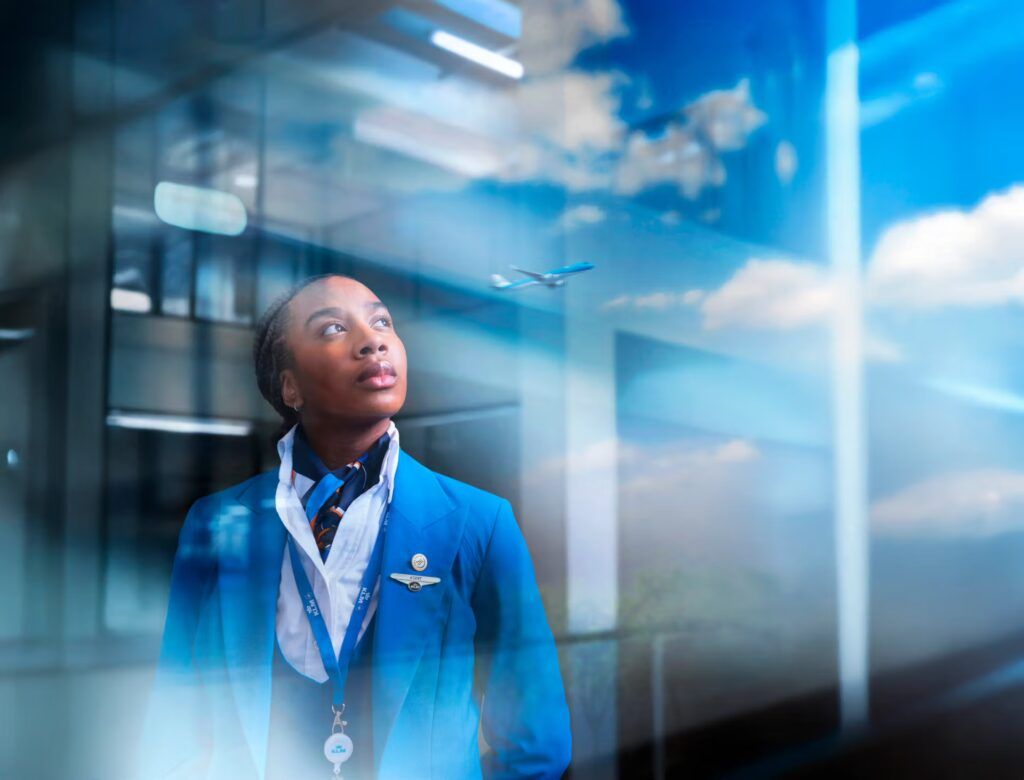 Een vrouw in een blauw uniform kijkt door een raam omhoog naar een vliegtuig in de lucht; heldere hemel, zon.