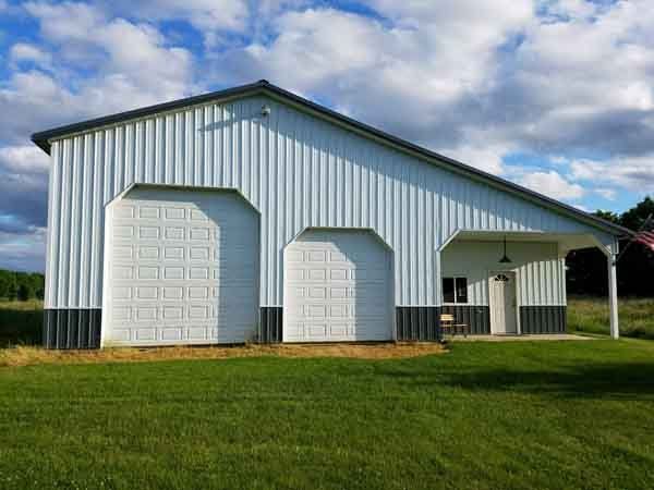 White metal barn with two garage doors and a porch, set in a grassy field under a cloudy sky.