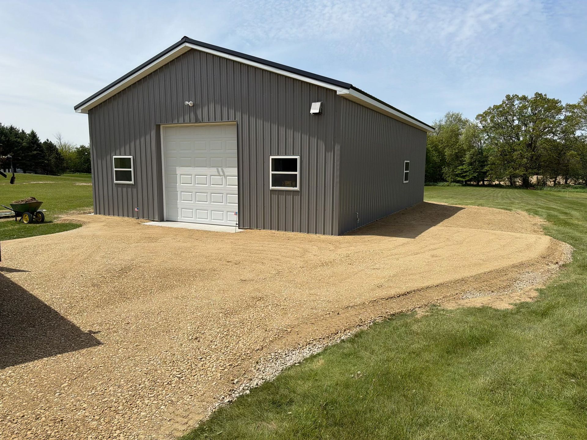 Gray metal barn with white garage door on a gravel driveway beside a grassy yard.