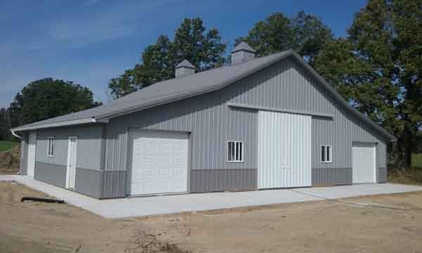 Gray and white metal barn with garage doors and a large sliding door, set on a concrete pad.