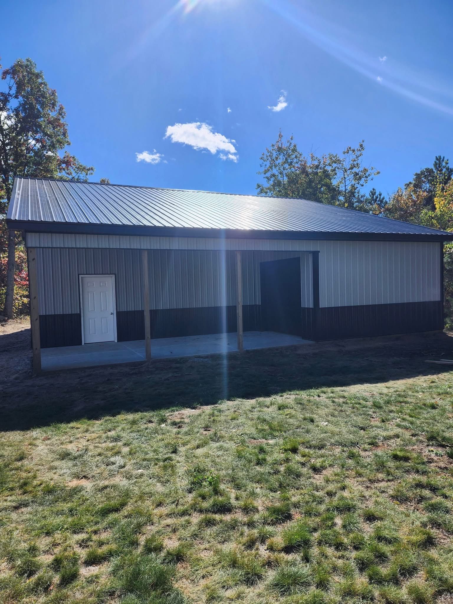Barn with a corrugated metal roof, gray and brown siding, and concrete patio, on a grassy plot under a blue sky.