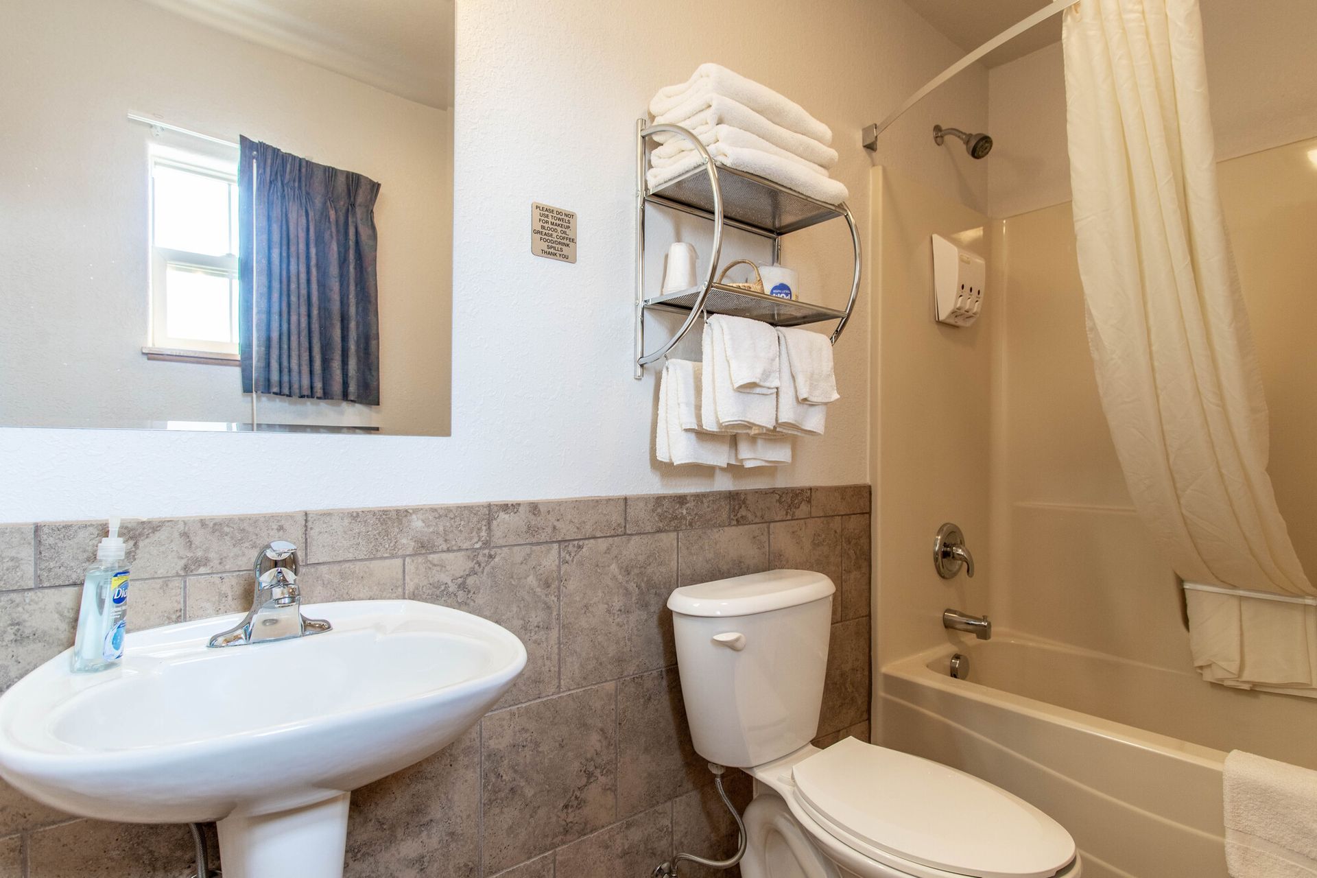 Bathroom with white sink, toilet, towels, and shower. Light walls and a window with blue curtains.