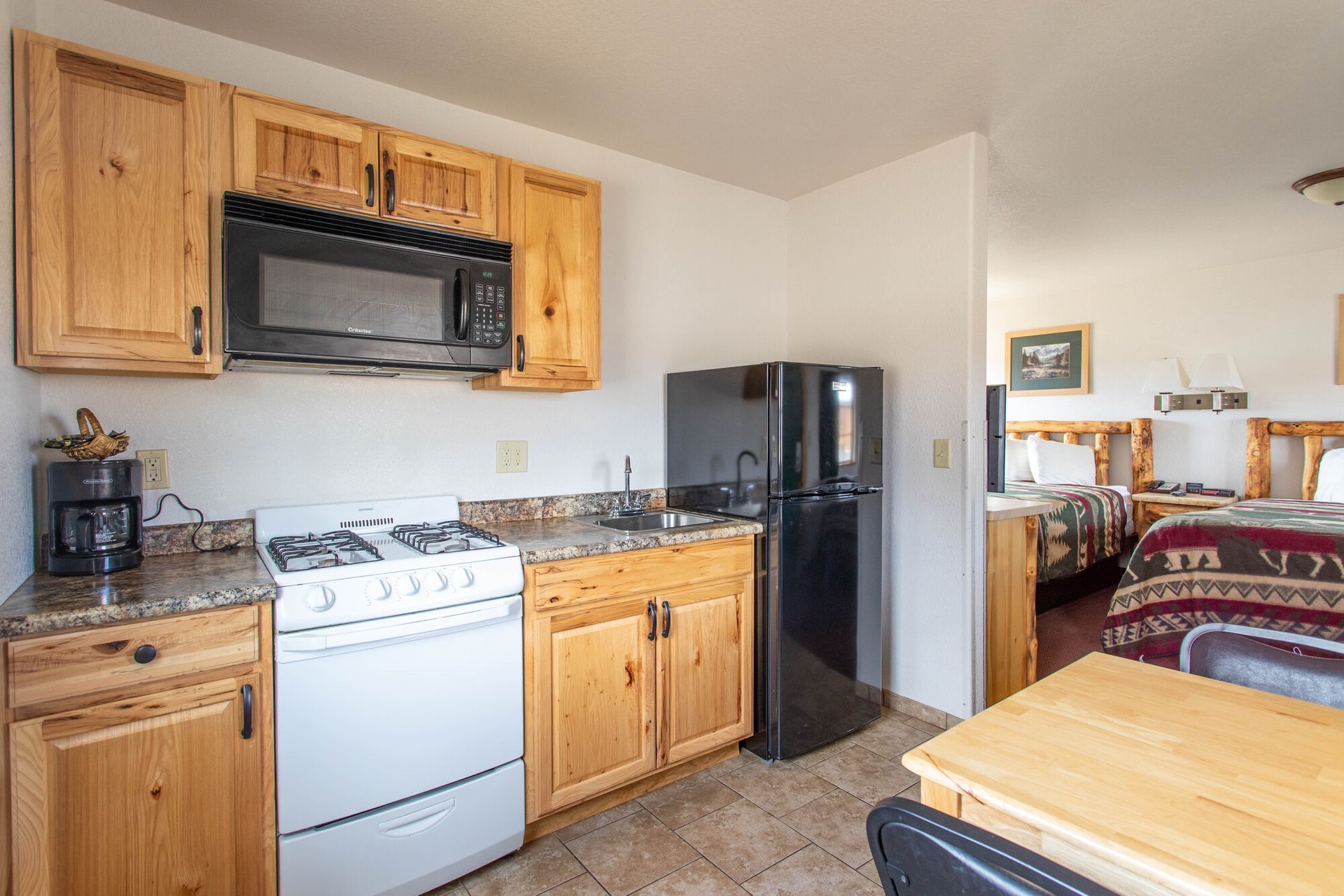 Small kitchen with stove, microwave, refrigerator, and cabinets, leading to a bedroom with beds.