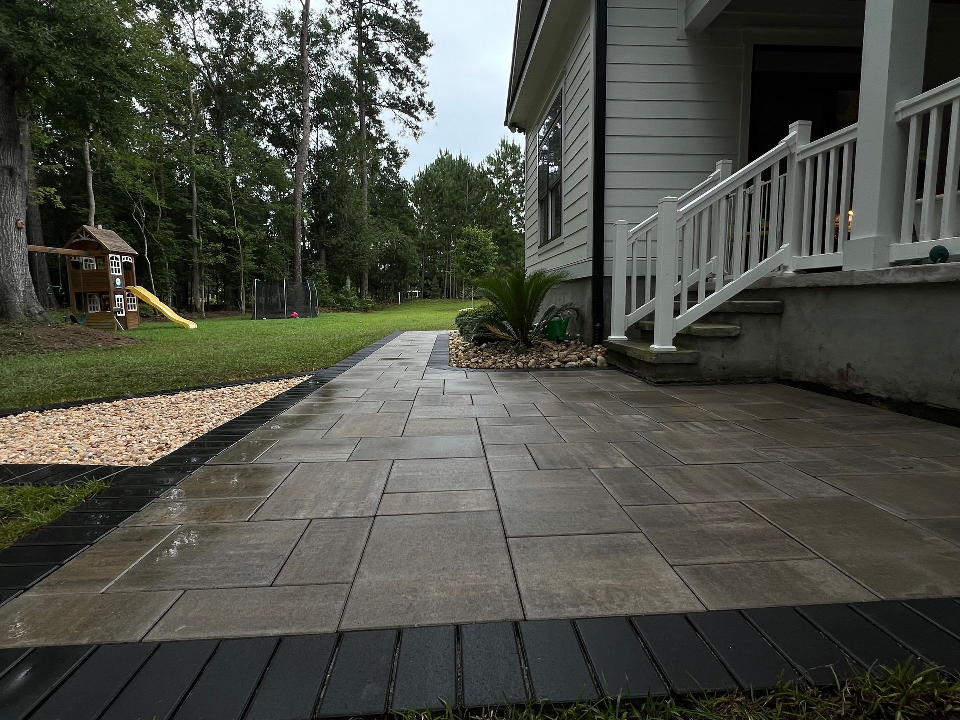 Stone patio walkway beside a house porch, with steps, railings, and trees in the yard.