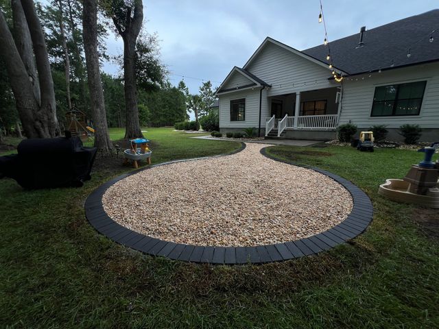 Circular stone fire pit in a grassy backyard near a white house and trees.