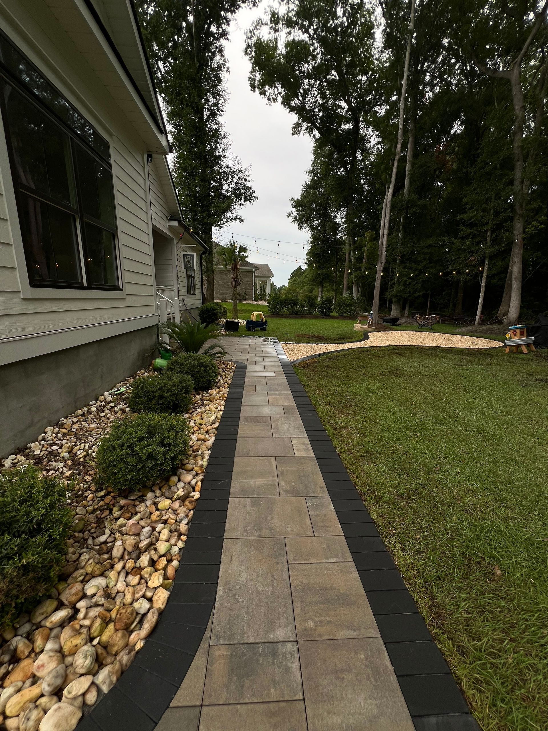 Stone walkway beside a house, bordered by landscaping, leading toward tall trees and a grassy yard.