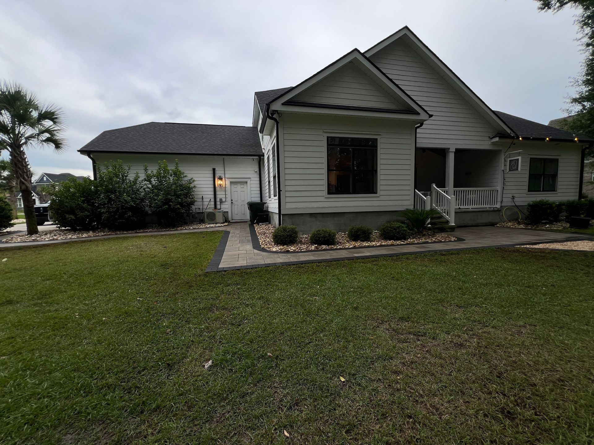 Front yard of a light gray house with a lawn, shrubs, and stone walkway under an overcast sky