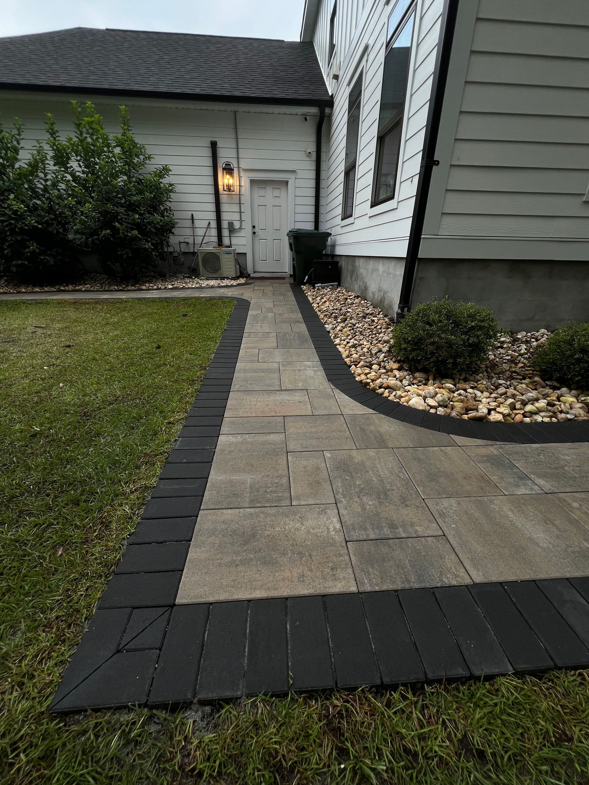 Front walkway with stone pavers leading to a house entry, bordered by dark bricks and landscaping stones.