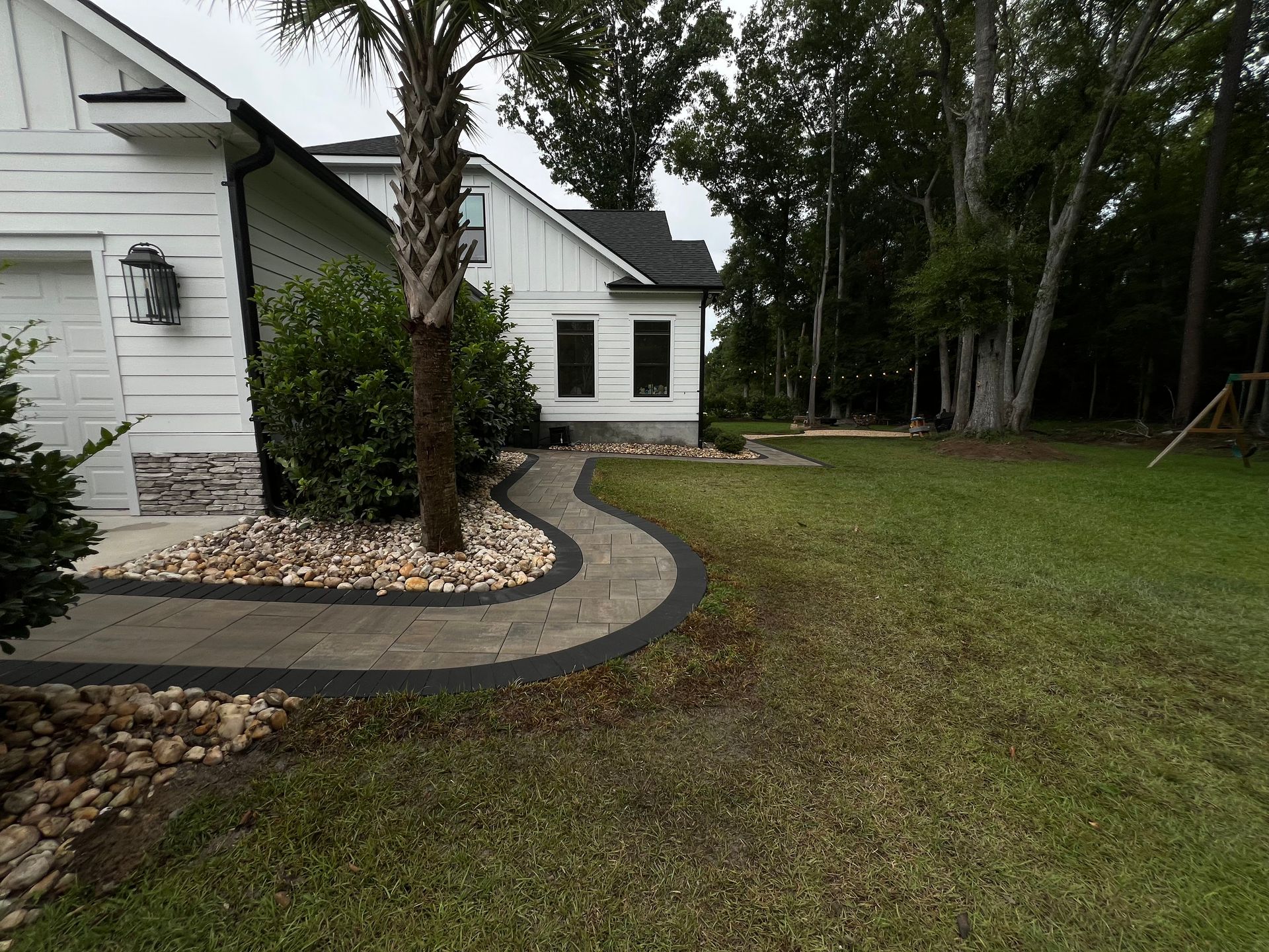 Curved stone walkway beside a white house, with a palm tree, rock border, and grassy yard.