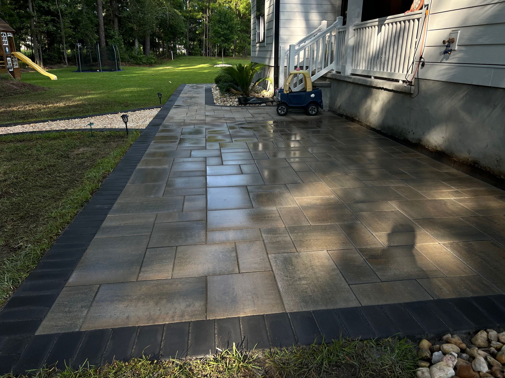 Wet stone patio beside a white house, reflecting sunlight, with a grassy yard and trees nearby.