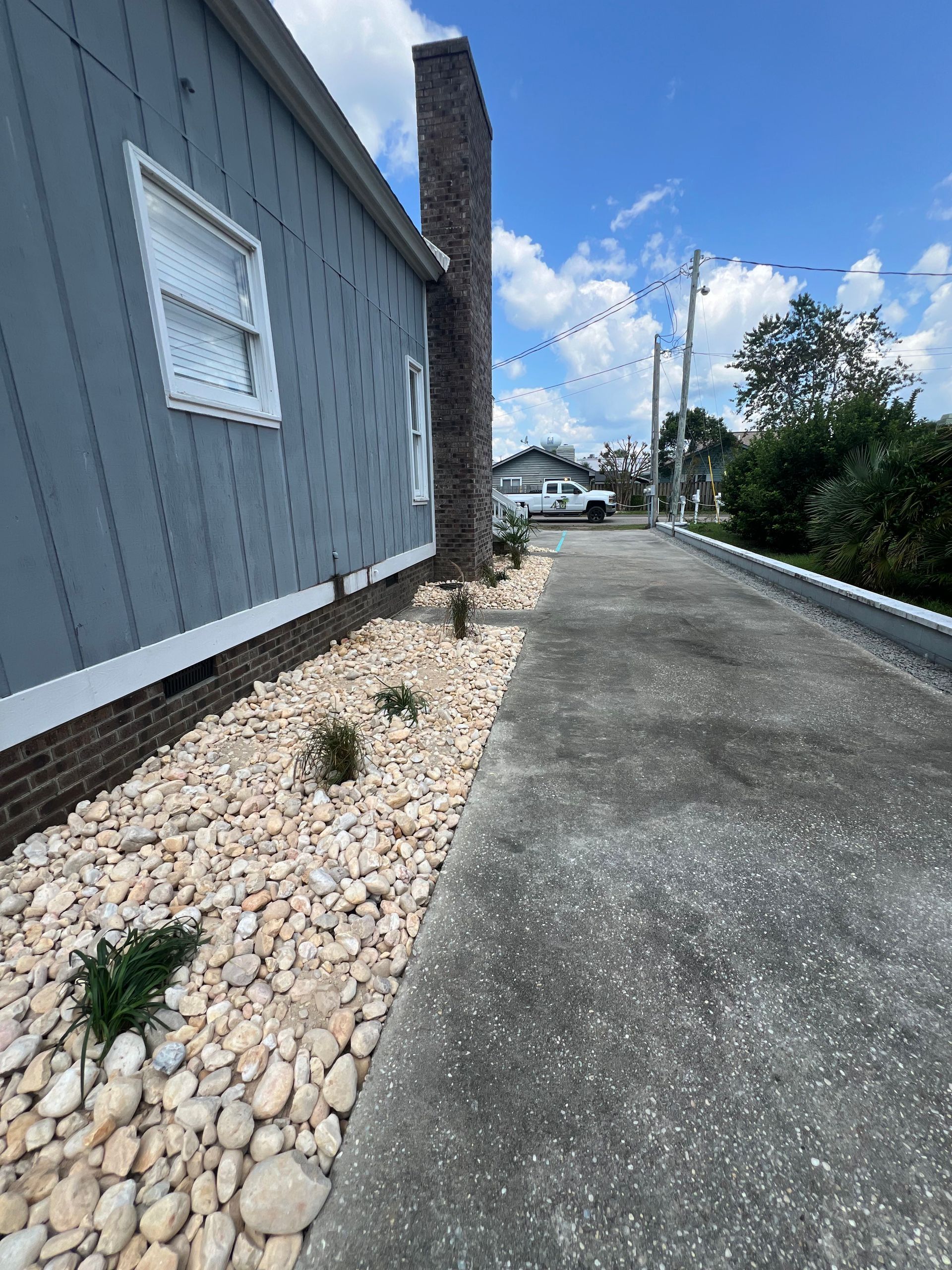 Narrow driveway beside a gray house, with white rock landscaping and a distant home under a blue sky.