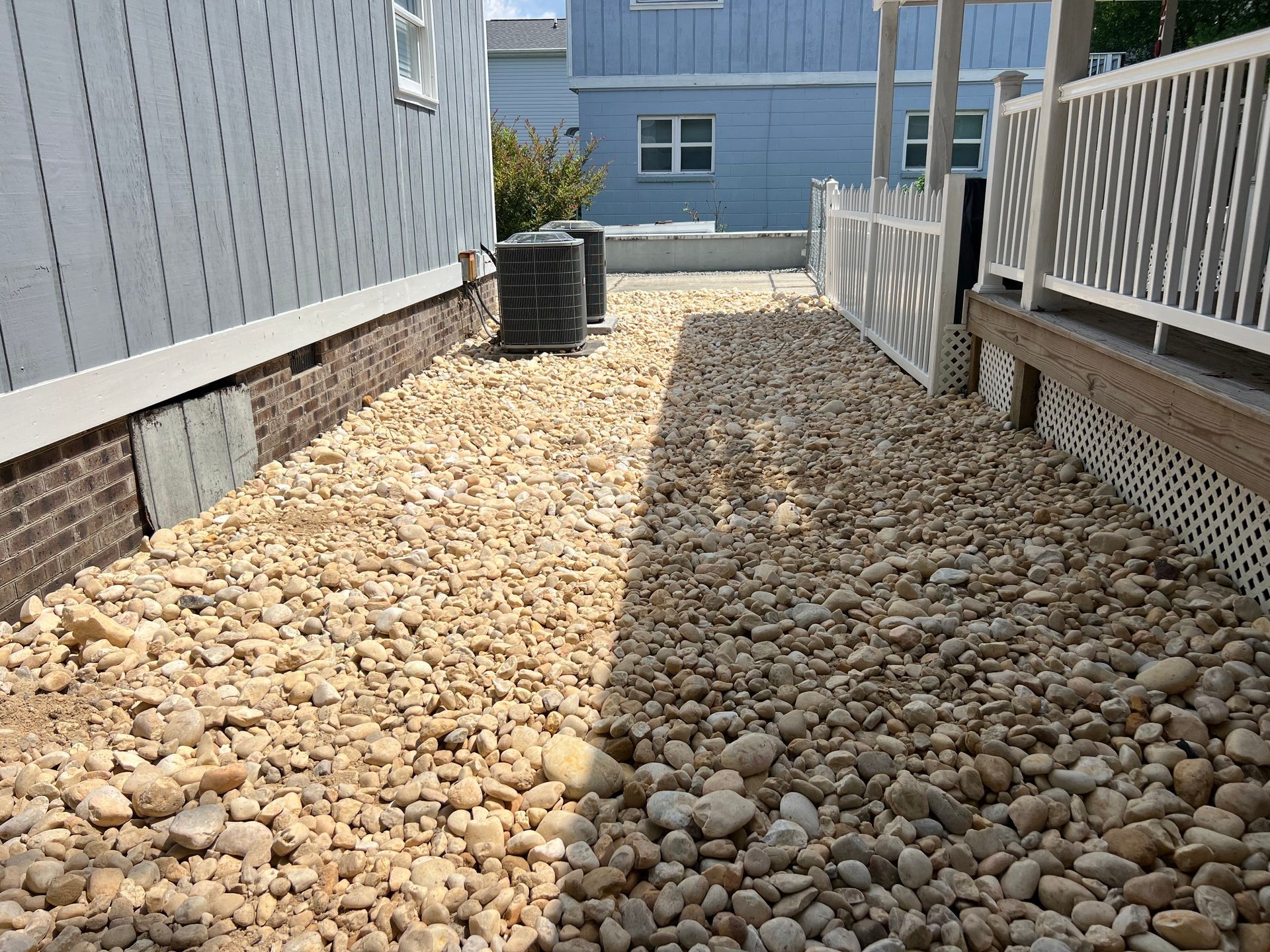 Rock-covered side yard between two buildings with a deck and railing on the right and a bin at the far end.