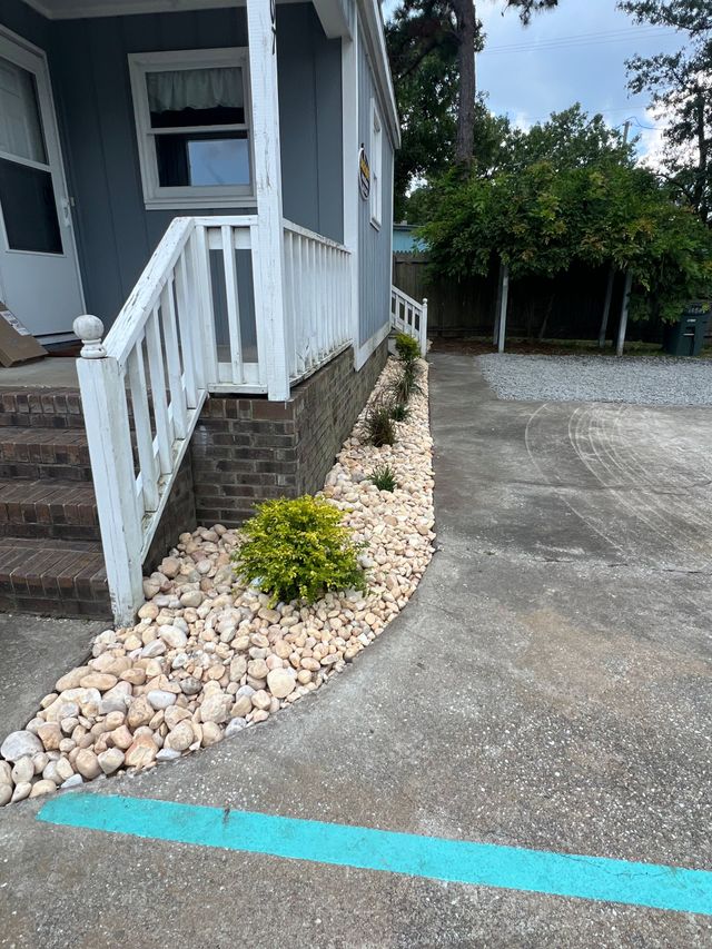 Front steps of a blue house with white railing, gravel driveway, and landscaped rock border with a small bush