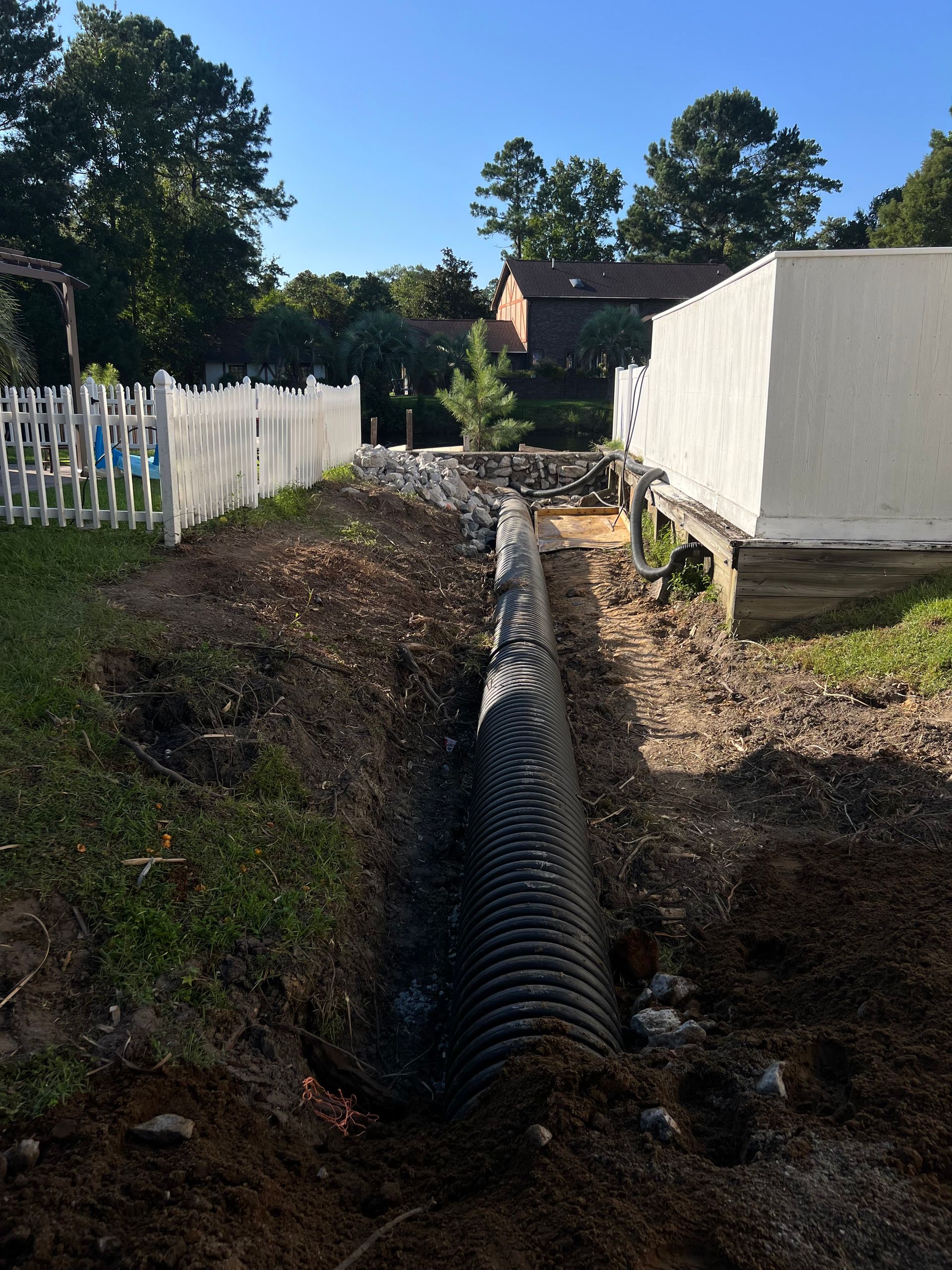 Dug trench with black drainage pipe beside a white fence and house in a backyard.