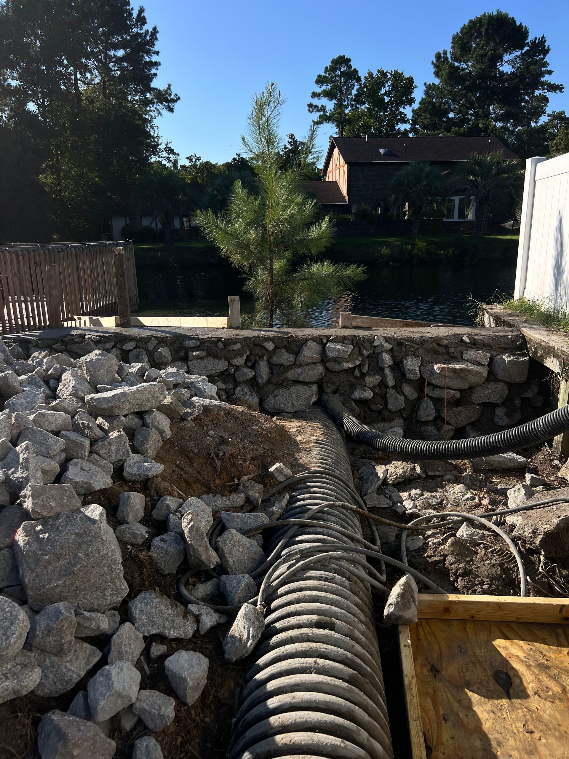 Rock-lined excavation site with a corrugated drain pipe, stone wall, and trees in the background