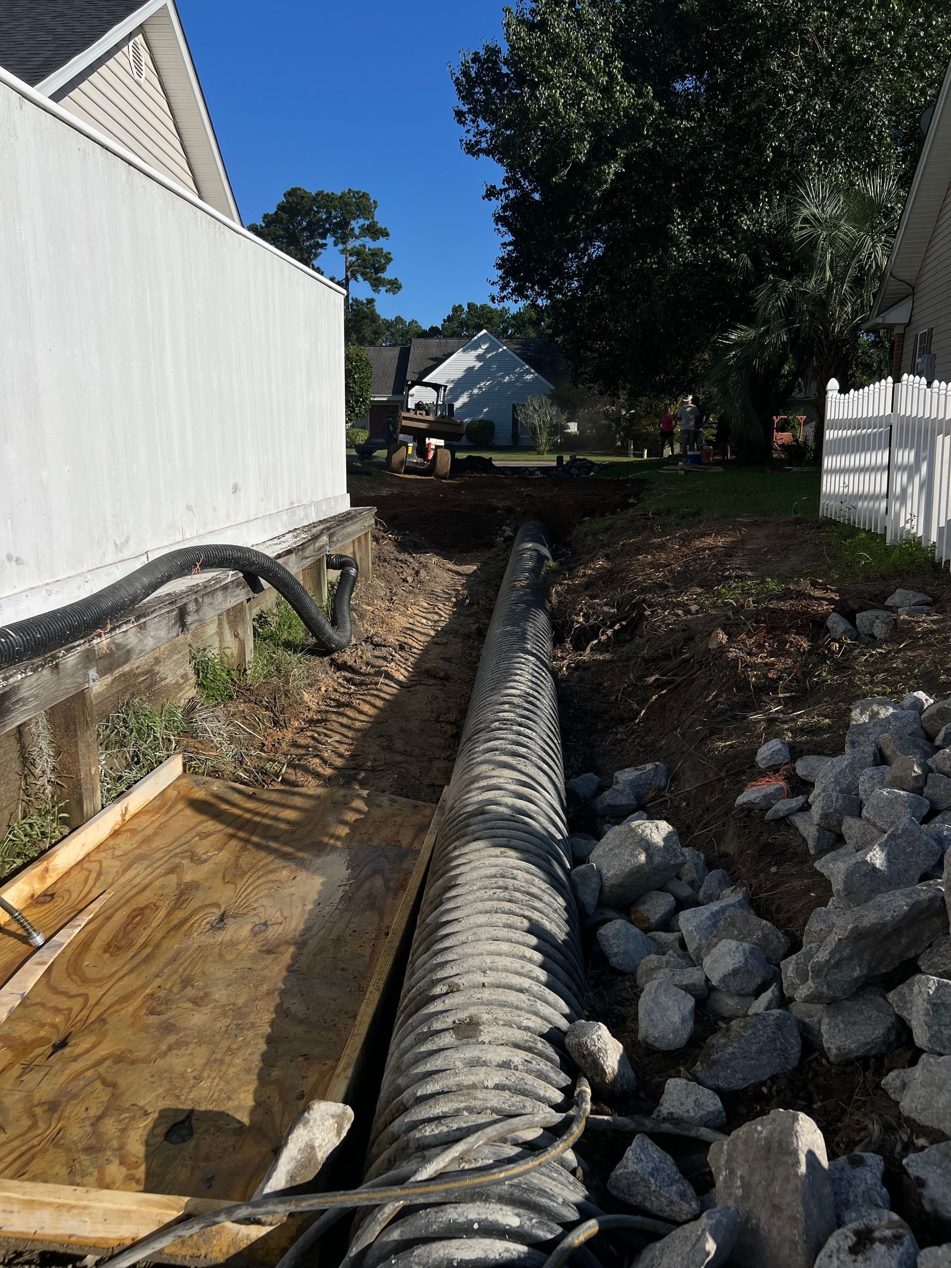 Backyard drainage trench with corrugated pipe, rocks, and dirt between white fences under blue sky