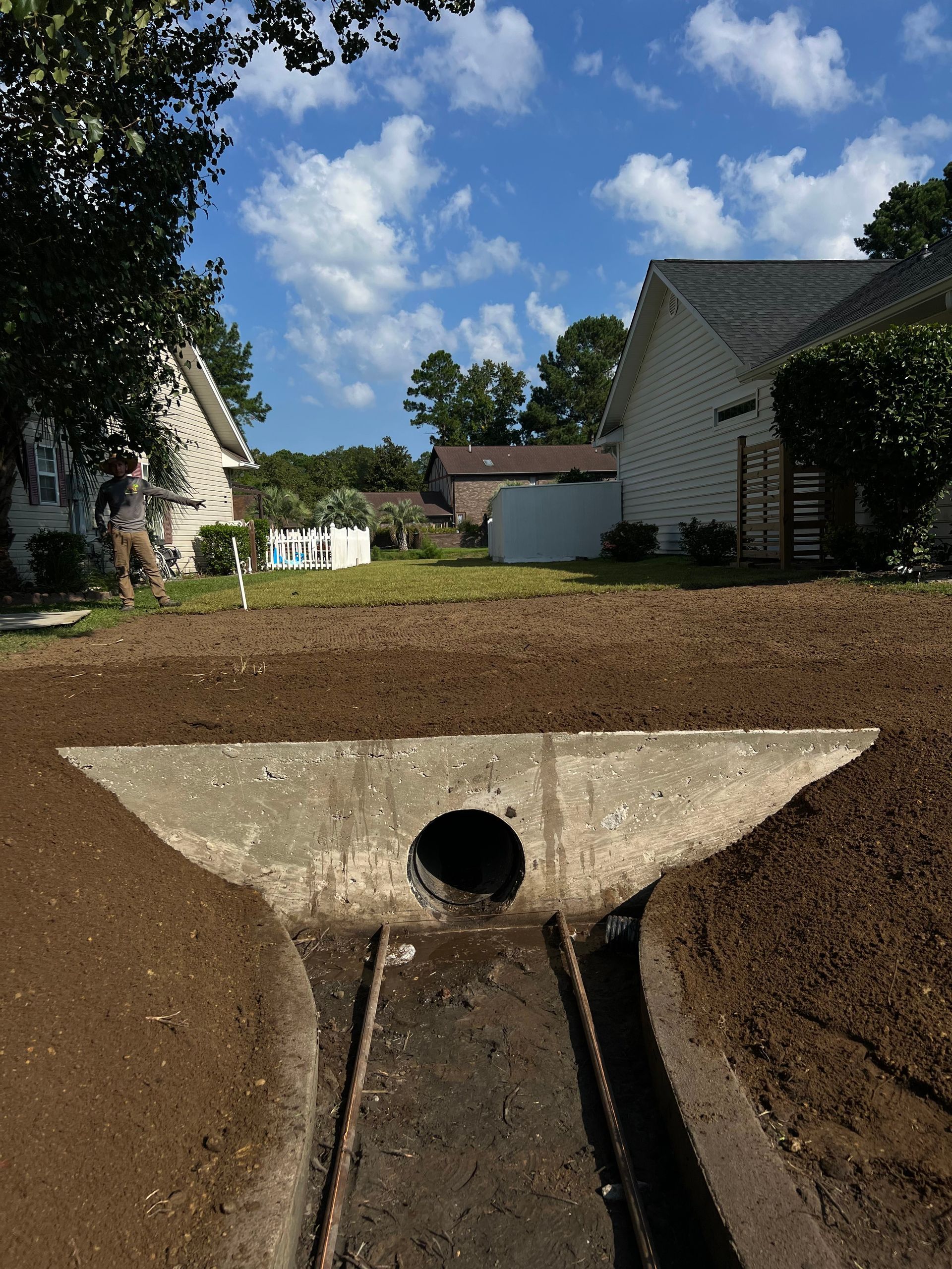 Open drainage culvert beneath a dirt slope, with homes and trees under a blue sky in the background