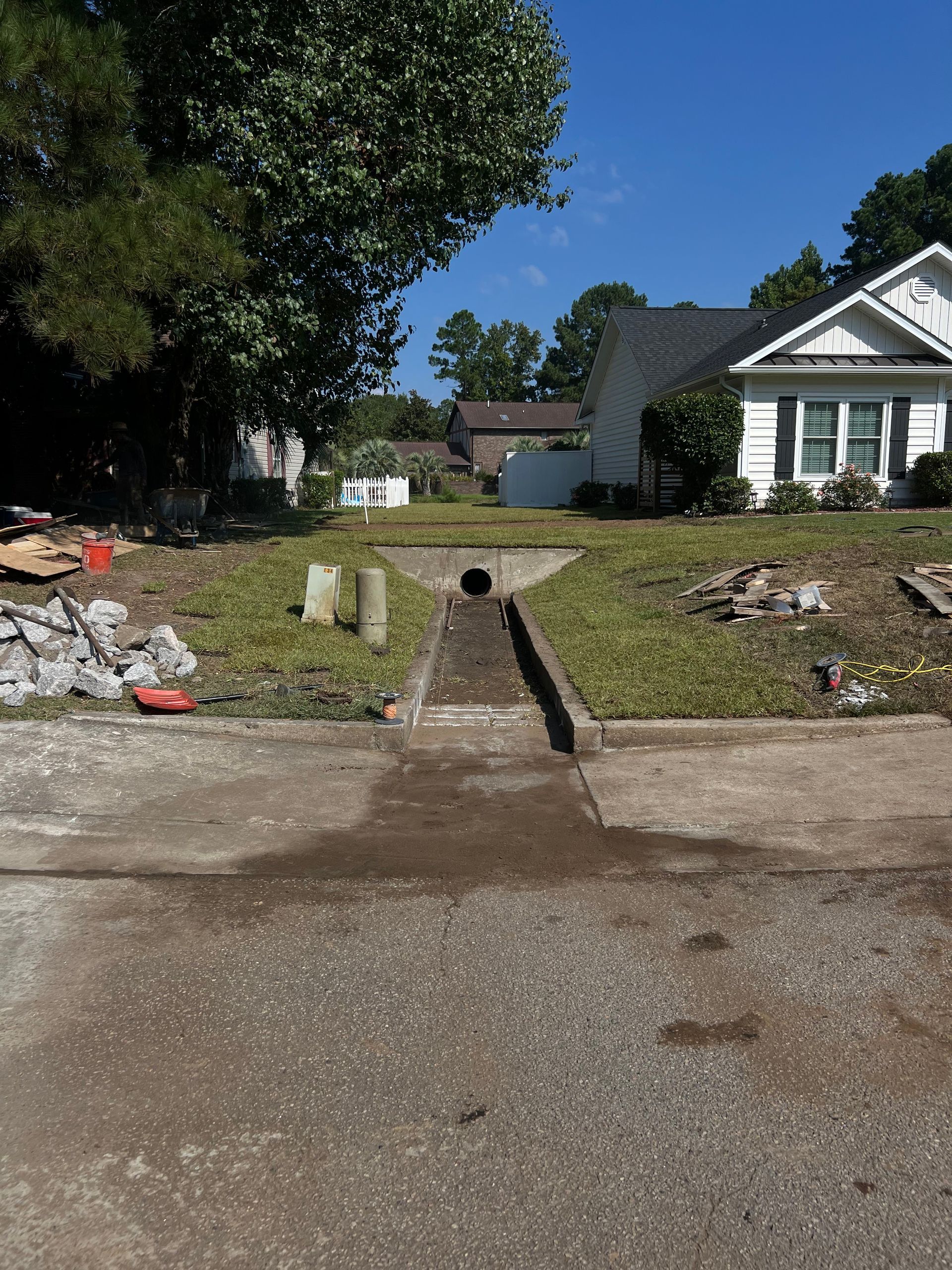 Damaged driveway and yard with debris, a broken retaining wall, and a white house under a clear blue sky