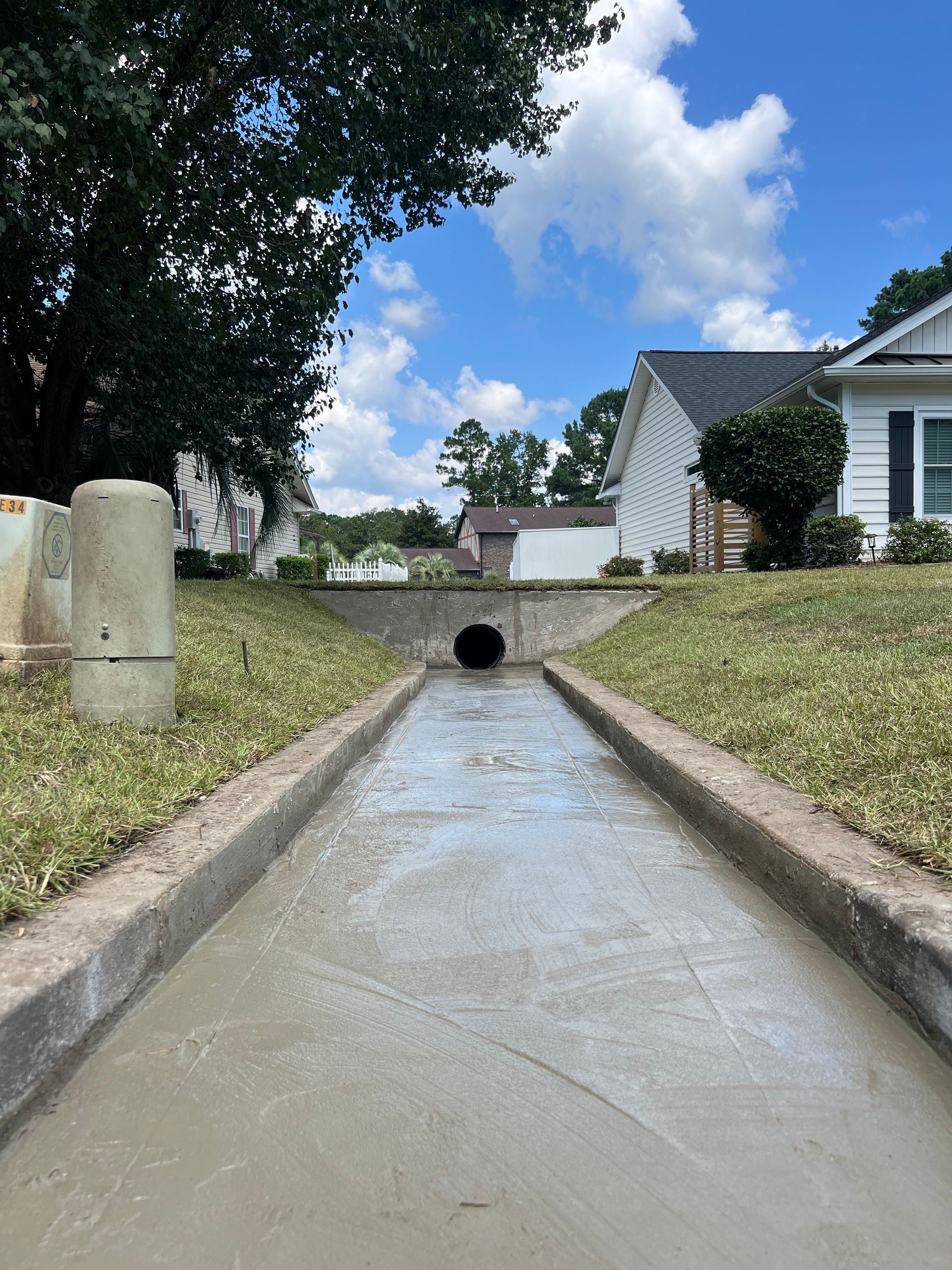 Concrete drainage ditch leading to a culvert under a road beside grassy yards and houses