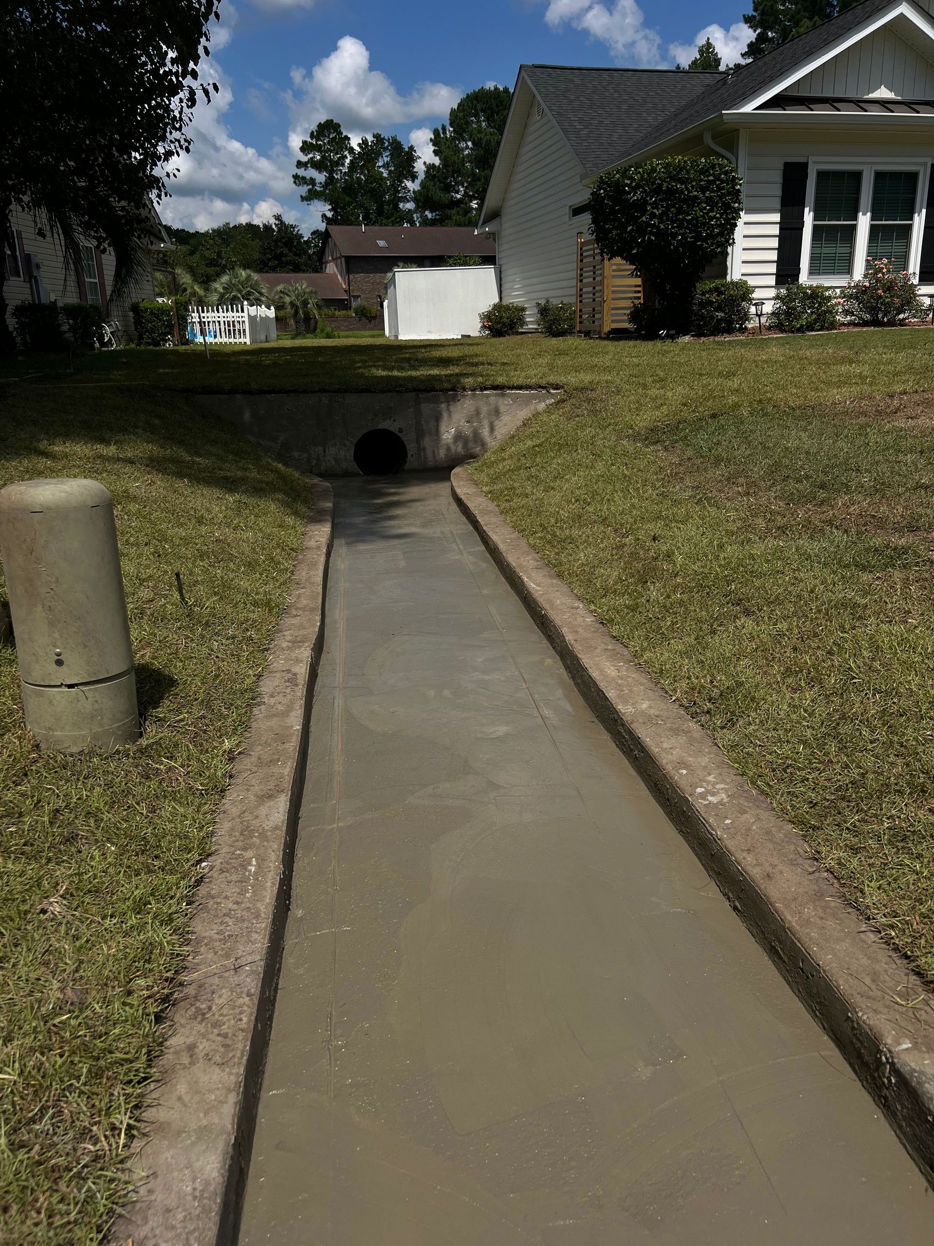 Concrete drainage channel leading to a culvert under a grassy residential slope, beside a house