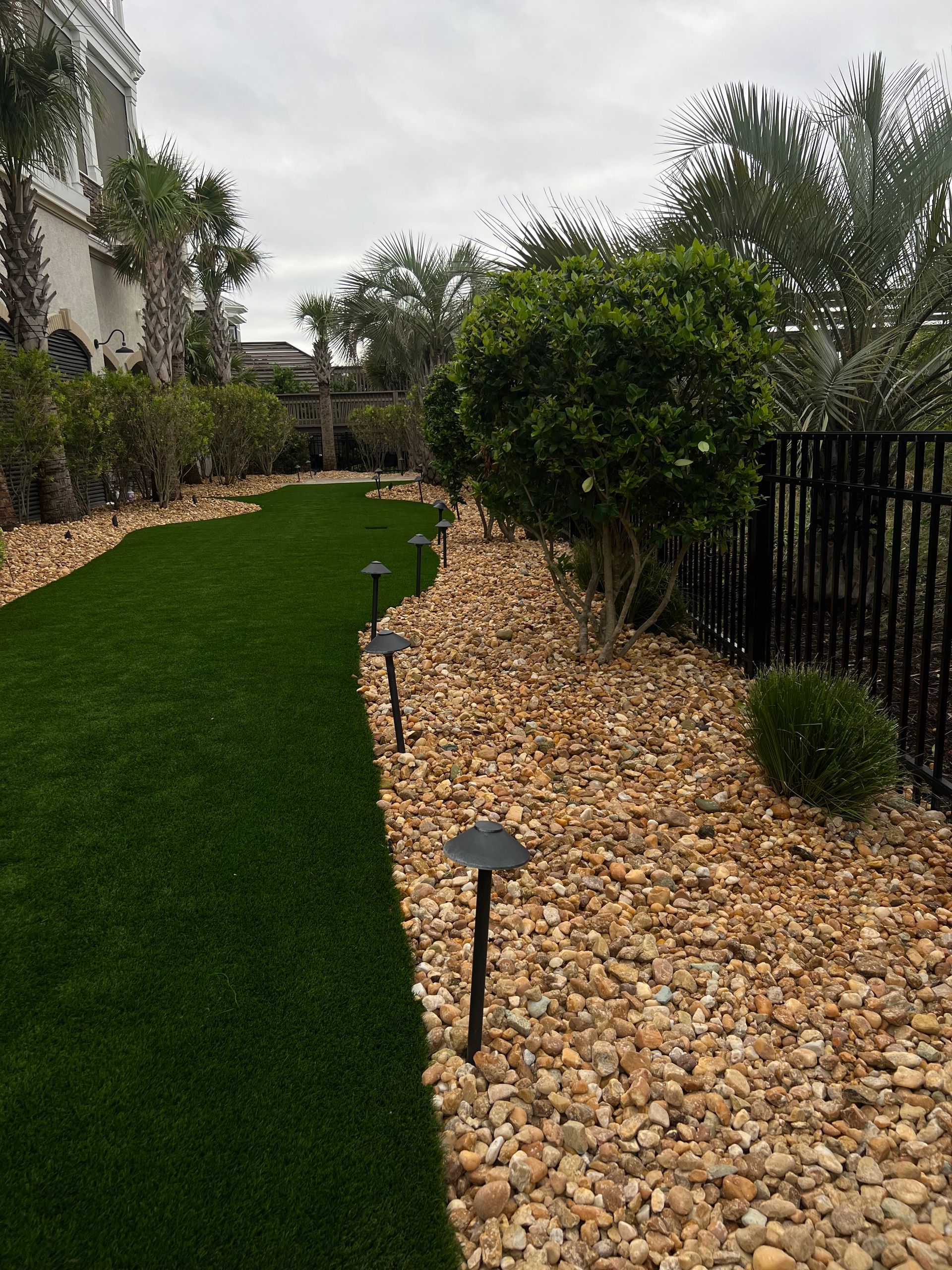Landscaped walkway with green turf, river rock border, and garden lights beside a fence and shrubs